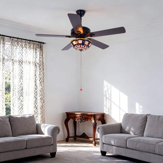 A dark bronze ceiling fan with five blades and a decorative stained-glass light fixture, installed in a bright, modern living room with two gray sofas and a wooden console table.